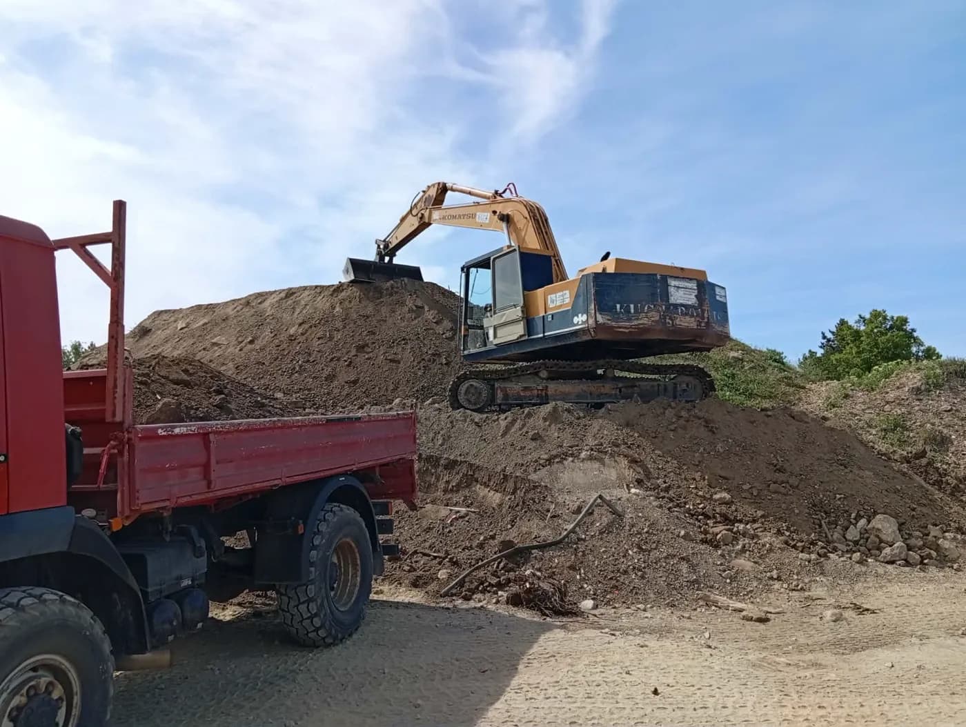 Komatsu lánctalpas kotró földhalmon, előtérben piros teherautó.